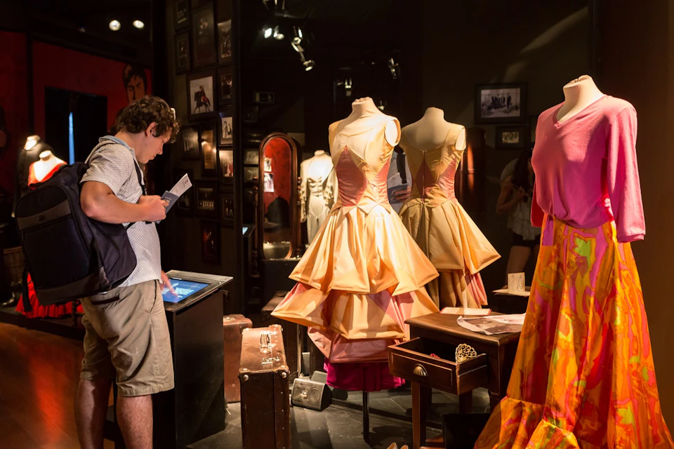 A person views an exhibit with mannequins displaying elaborate, layered dresses. Surrounding walls feature framed pictures and mirrors.
