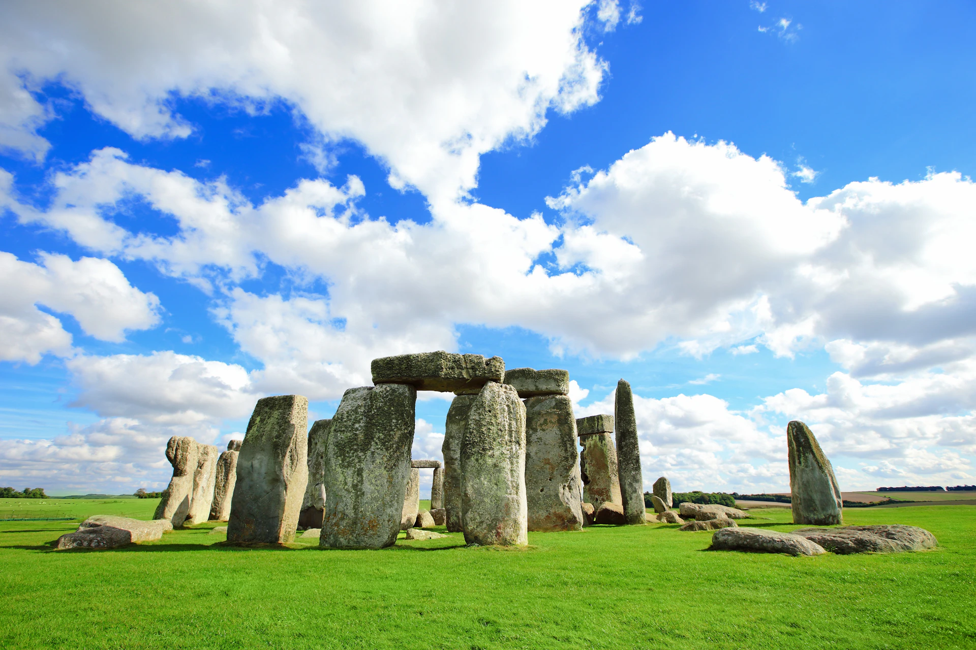 Stonehenge under a partly cloudy blue sky, surrounded by green grass.
