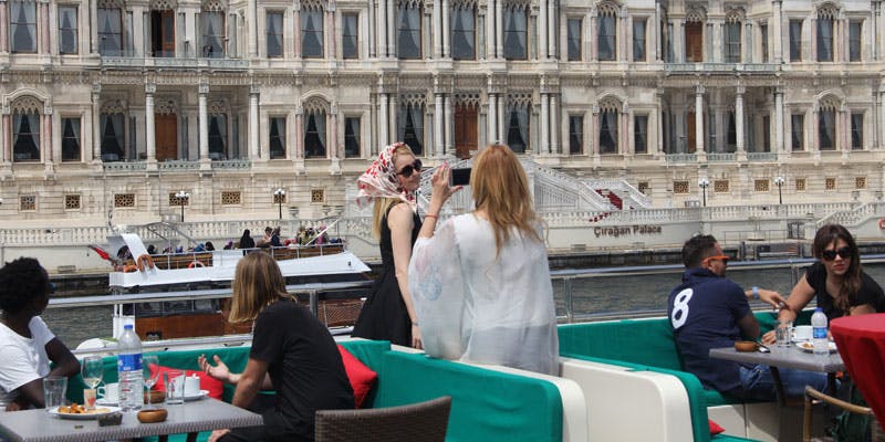 Three people on a boat near an ornate building. One woman is taking a photo, while a man and another woman sit at a table.