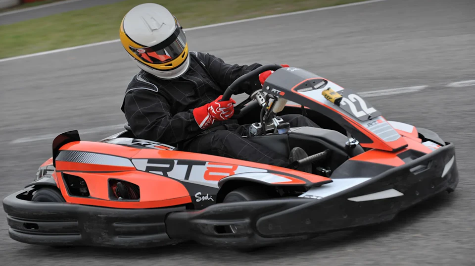 Person in a racing suit and helmet driving an orange and black go-kart on a track.