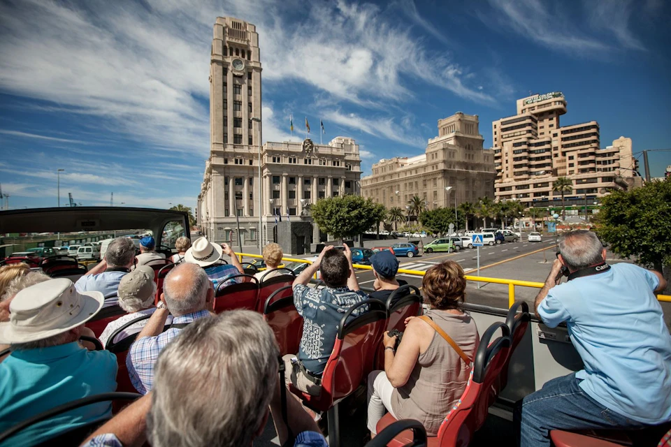 People seated on an open-top tour bus, taking photos of large historic buildings under a partly cloudy blue sky.