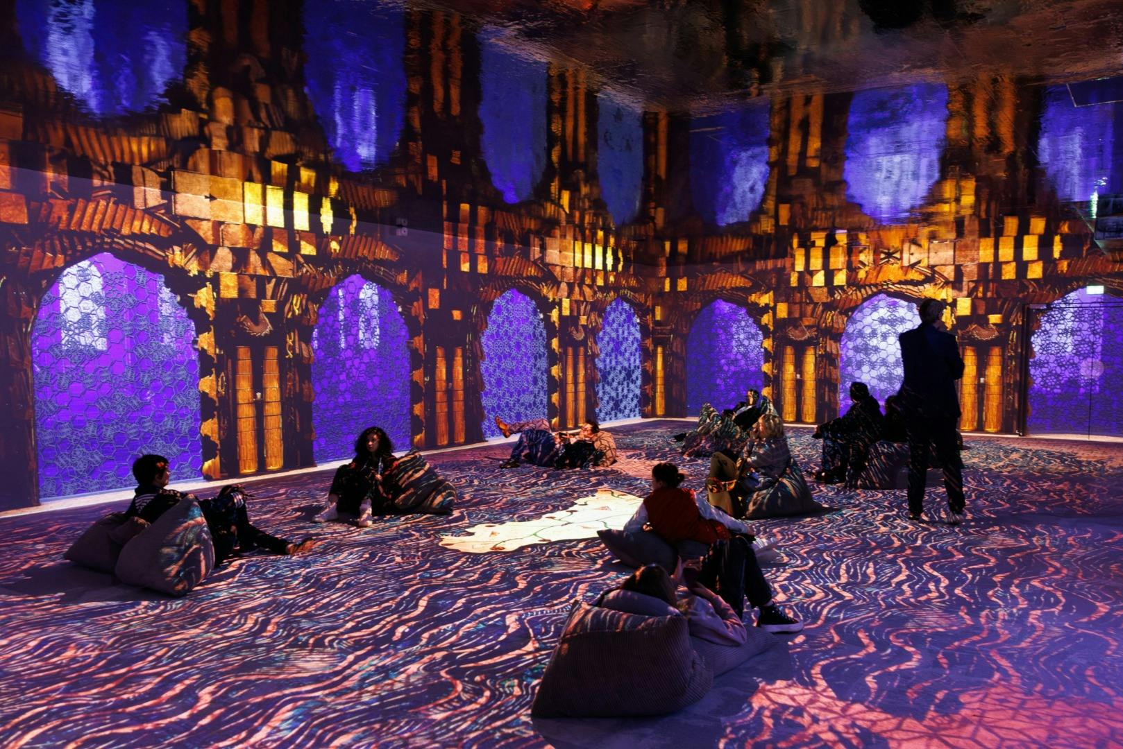 Persones assegudes o de peu en una habitació immersiva amb patrons de colors projectats a les parets i al terra.