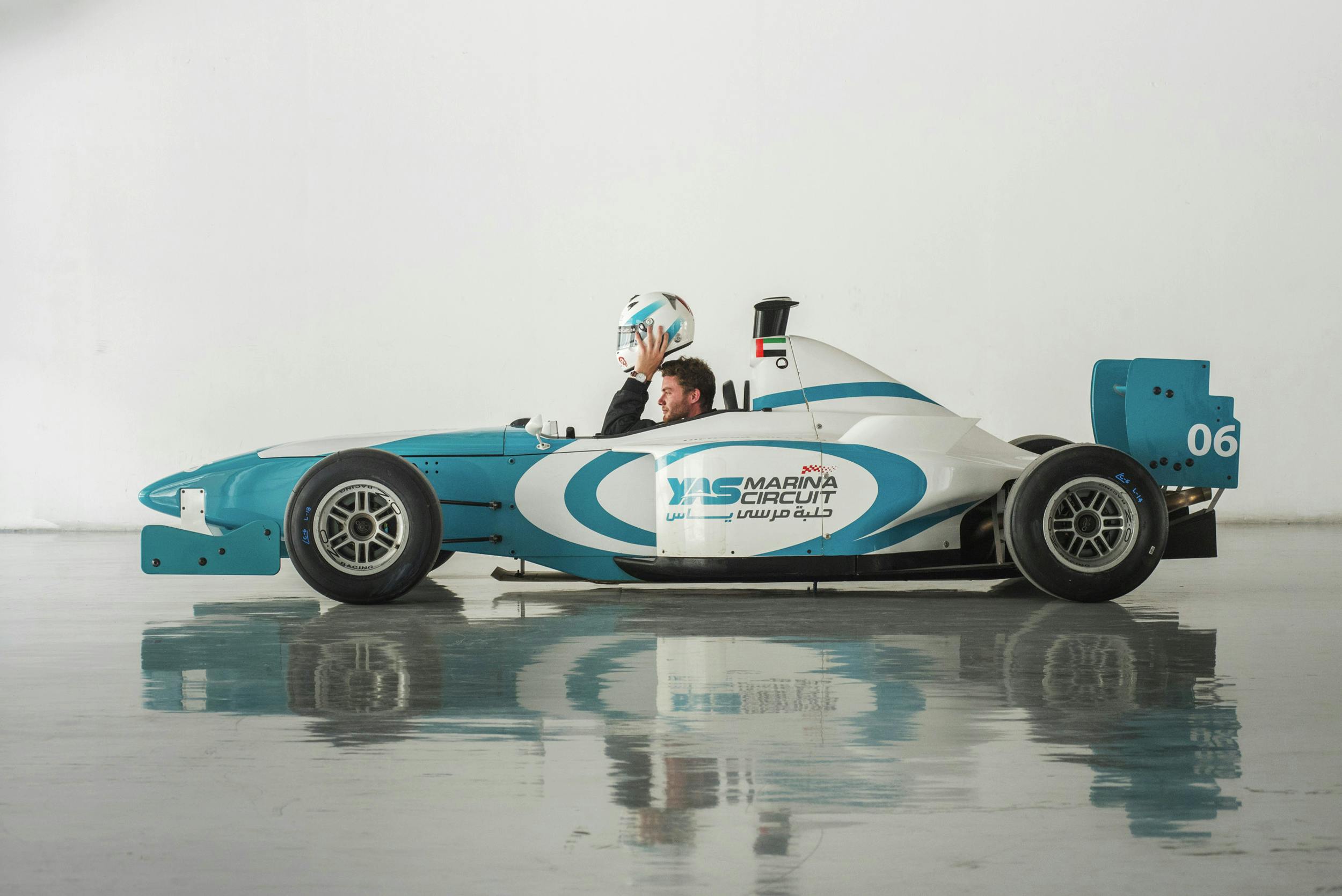 A man sitting in a blue and white race car on a reflective surface, holding a helmet. The car displays "Yas Marina Circuit" branding.