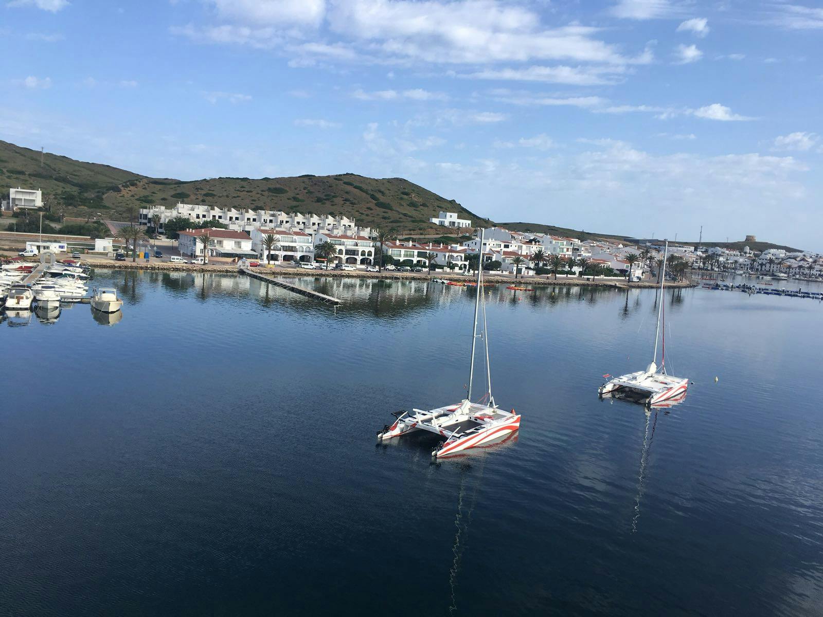 Two catamarans are docked in calm water, with a coastal town and hills in the background under a partly cloudy sky.