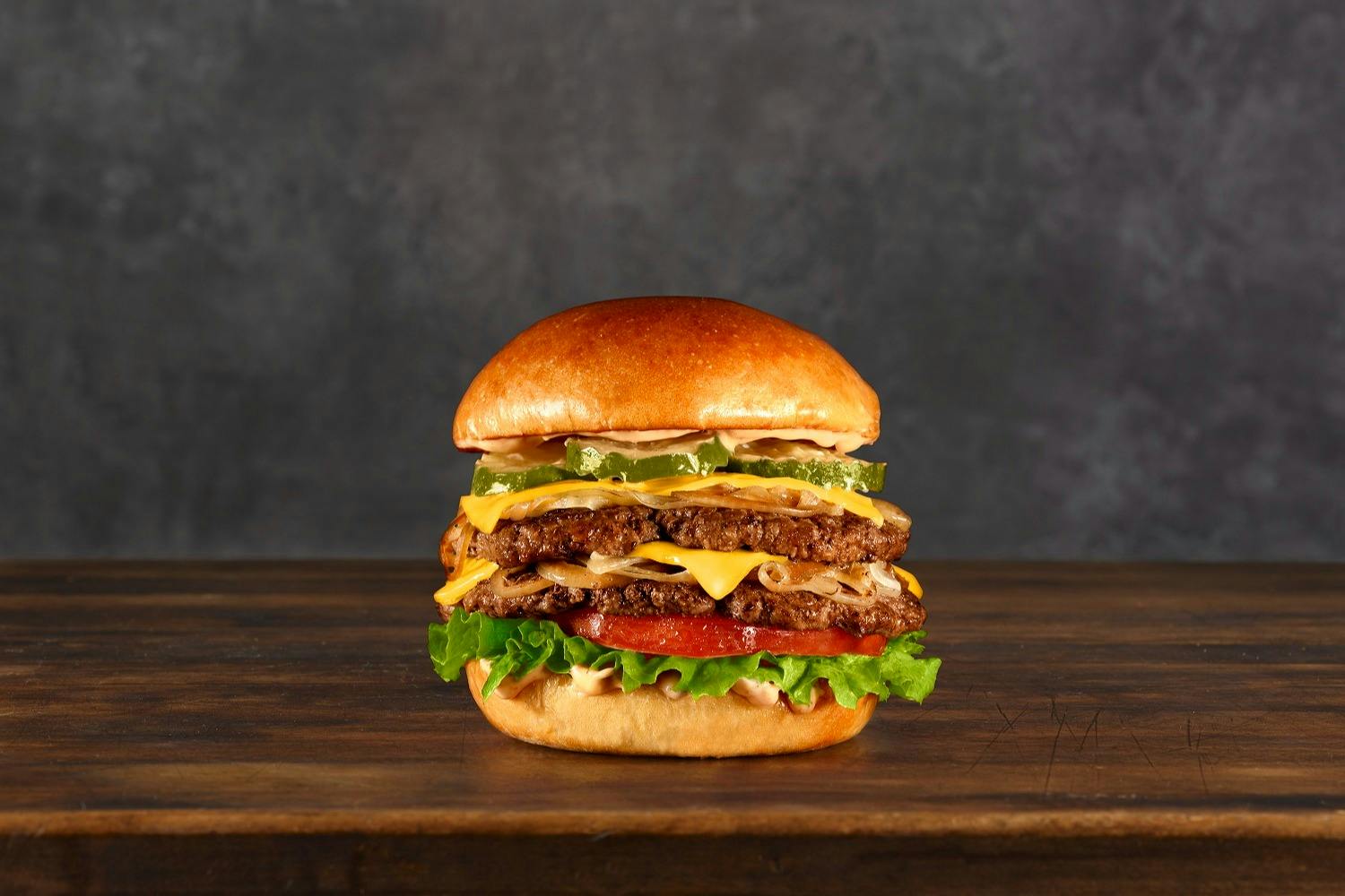 Ένα στοιβαγμένο cheeseburger με μαρούλι, ντομάτα, τυρί και πίκλες σε ξύλινη επιφάνεια σε σκούρο φόντο.