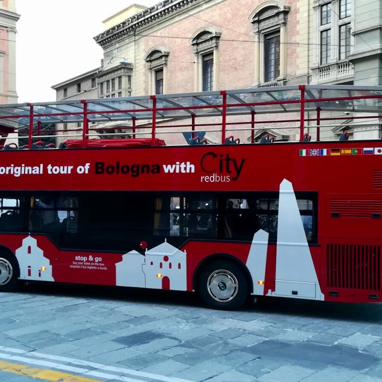 A red double-decker tour bus in Bologna with architectural graphics and multilingual text promoting city tours.
