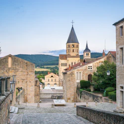 Cobblestone street leading to historic buildings and church towers, with hills and greenery in the background under a blue sky.