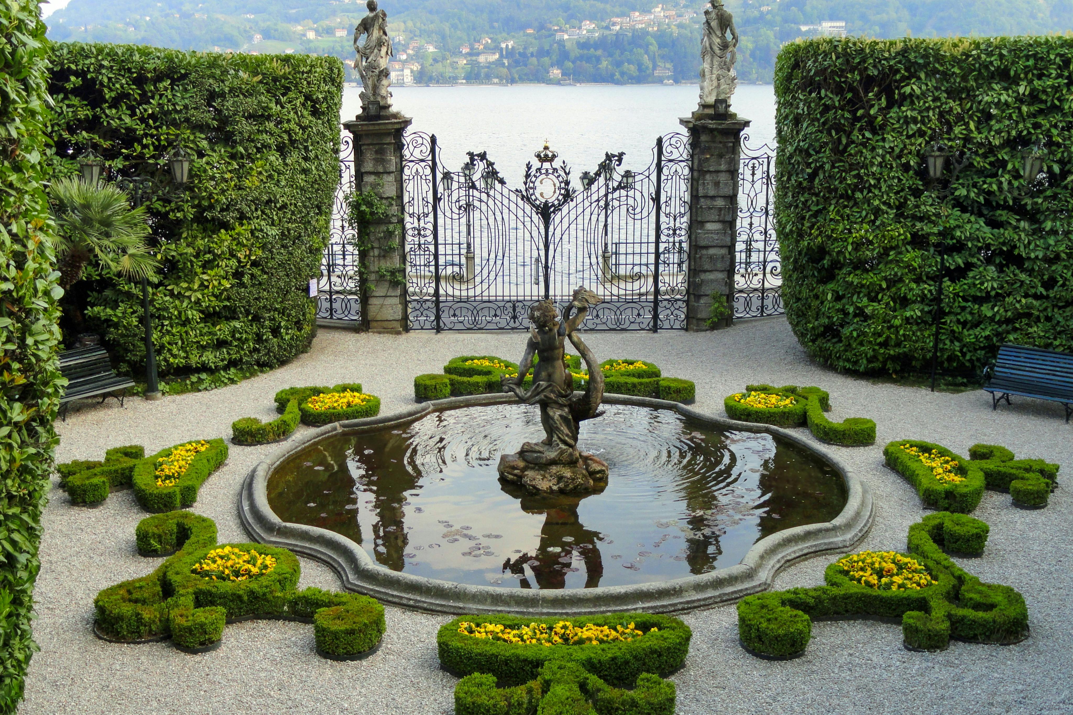 Decorative fountain with a statue in a formal garden, surrounded by hedges and overlooking a lake with ornate gates.