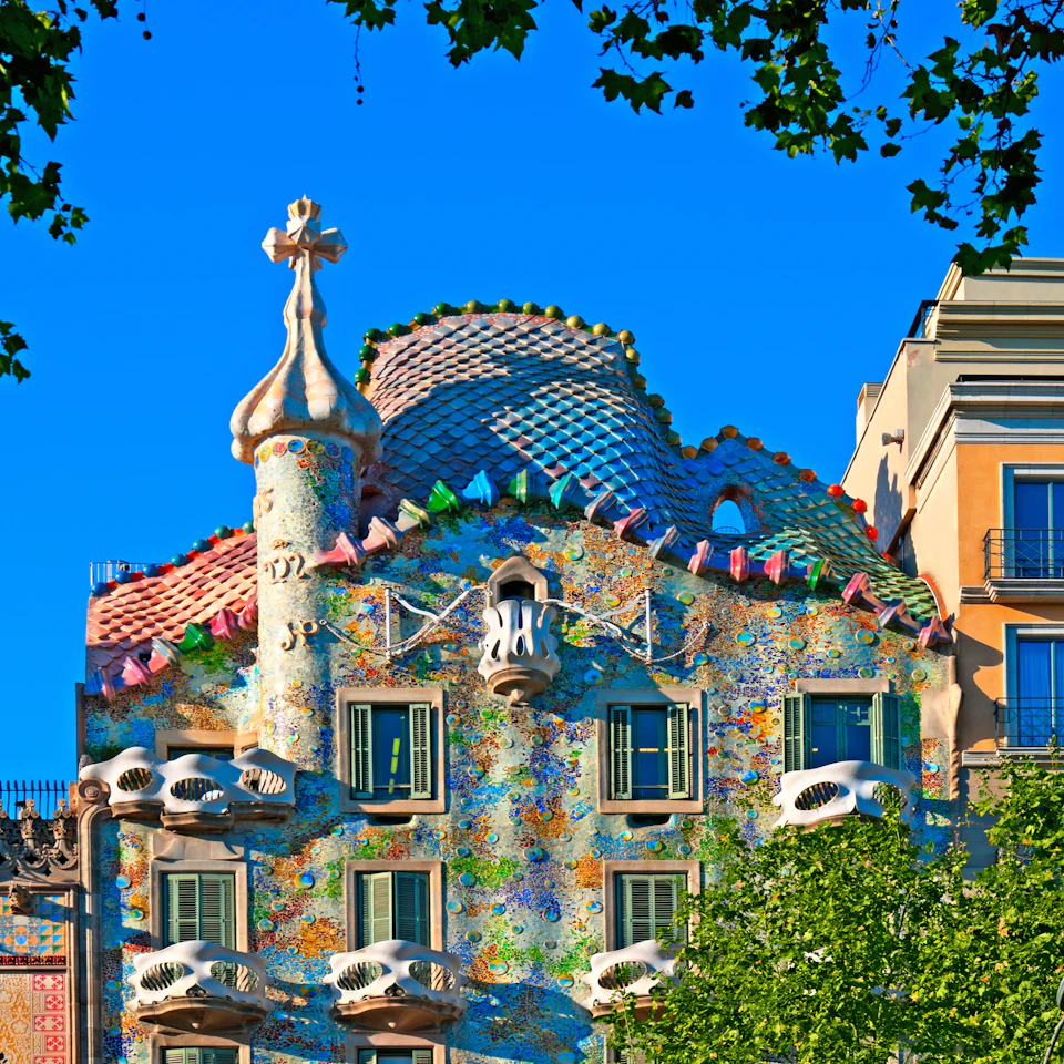 Colorful, ornate building with mosaic tiles, curved lines, and organic shapes under a bright blue sky, surrounded by green trees.