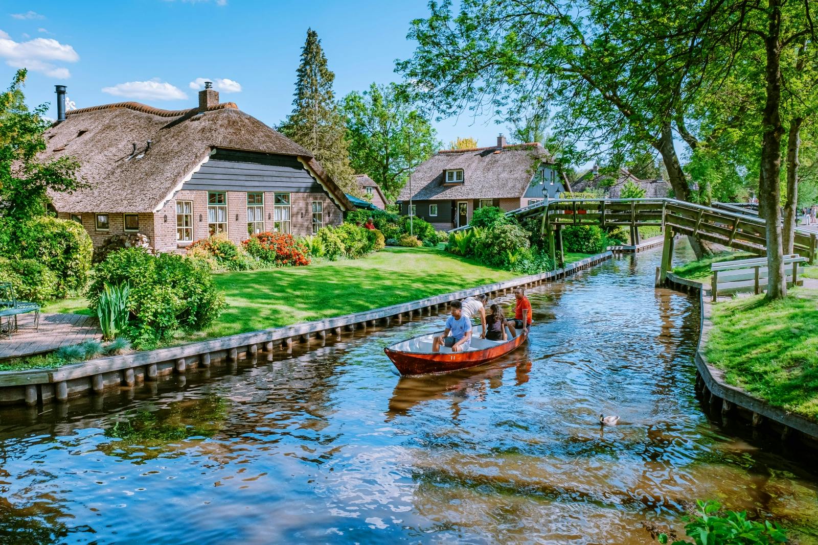 A scenic canal with a small wooden boat carrying four people, beside picturesque houses with thatched roofs and a wooden bridge.