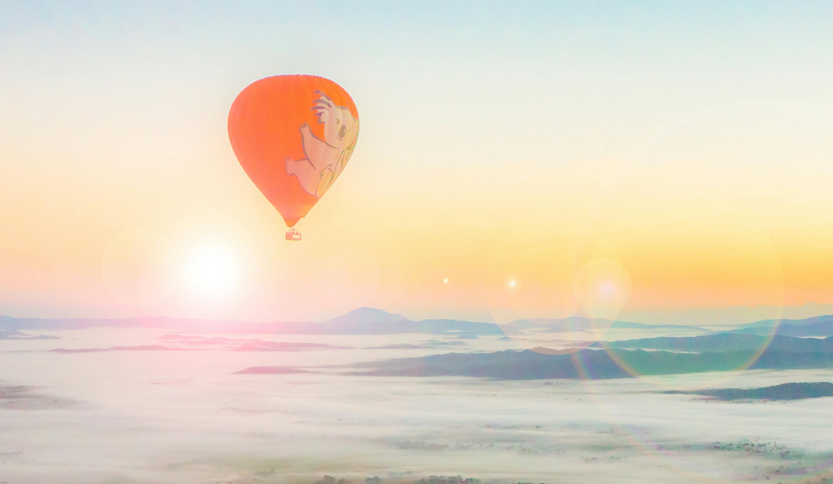 A vibrant orange hot air balloon with cartoon character ascends over a misty, mountainous landscape at sunrise.