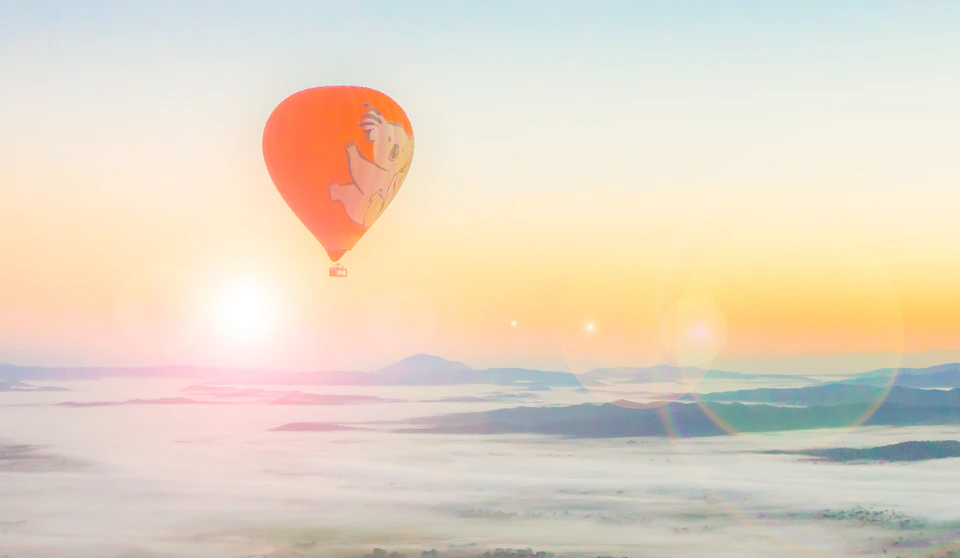 A vibrant orange hot air balloon with cartoon character ascends over a misty, mountainous landscape at sunrise.