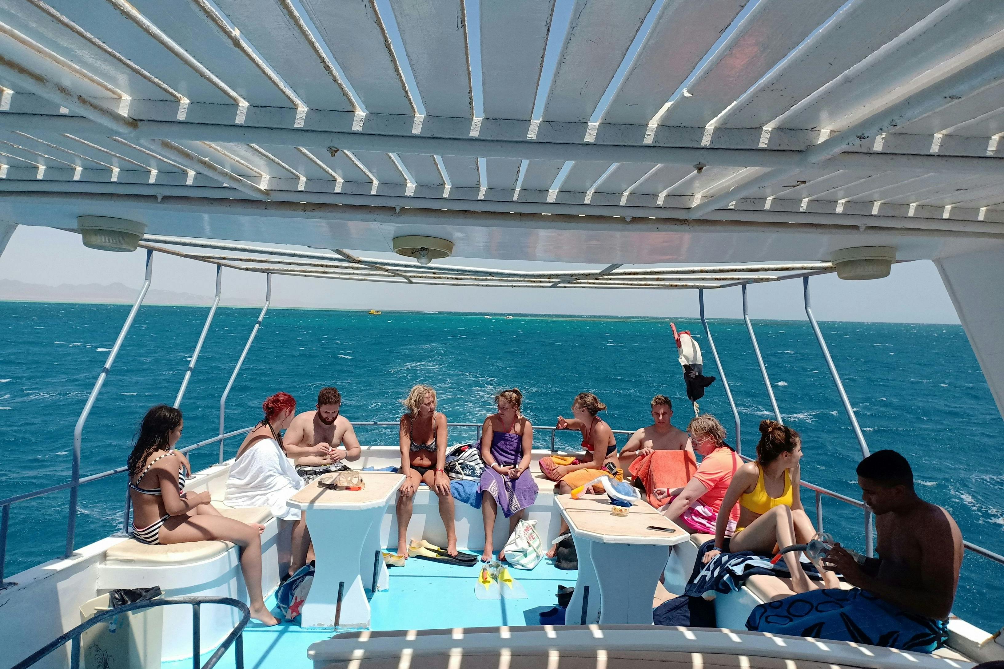 En gruppe mennesker i badetøj sidder og sludrer under en skyggefuld baldakin på en båd, med havet synligt i baggrunden.