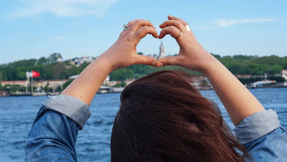 Person facing water, forming a heart shape with their hands. Background includes a hill with buildings and a tower.