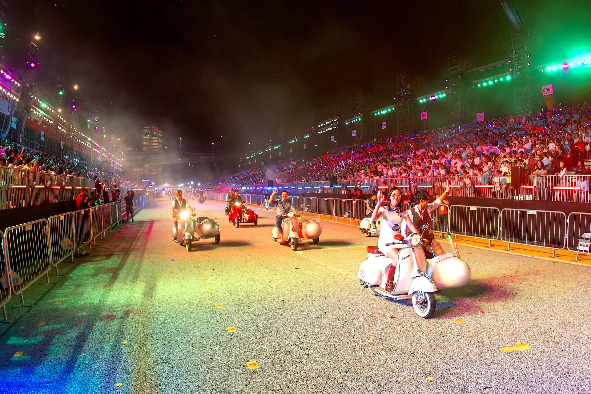 Rows of people on motor scooters ride down a street at night; large stands filled with spectators are seen in the background.