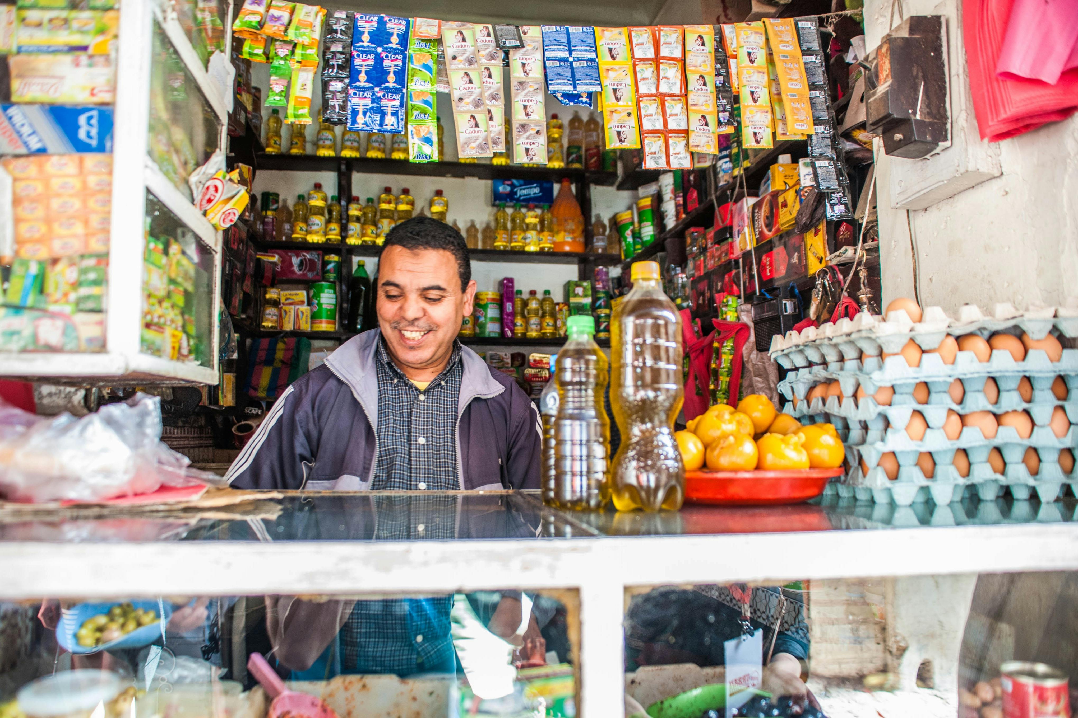 一个男人站在一家小商店的柜台后面，里面摆满了各种包装商品、蔬菜和瓶子，心满意足地微笑着。