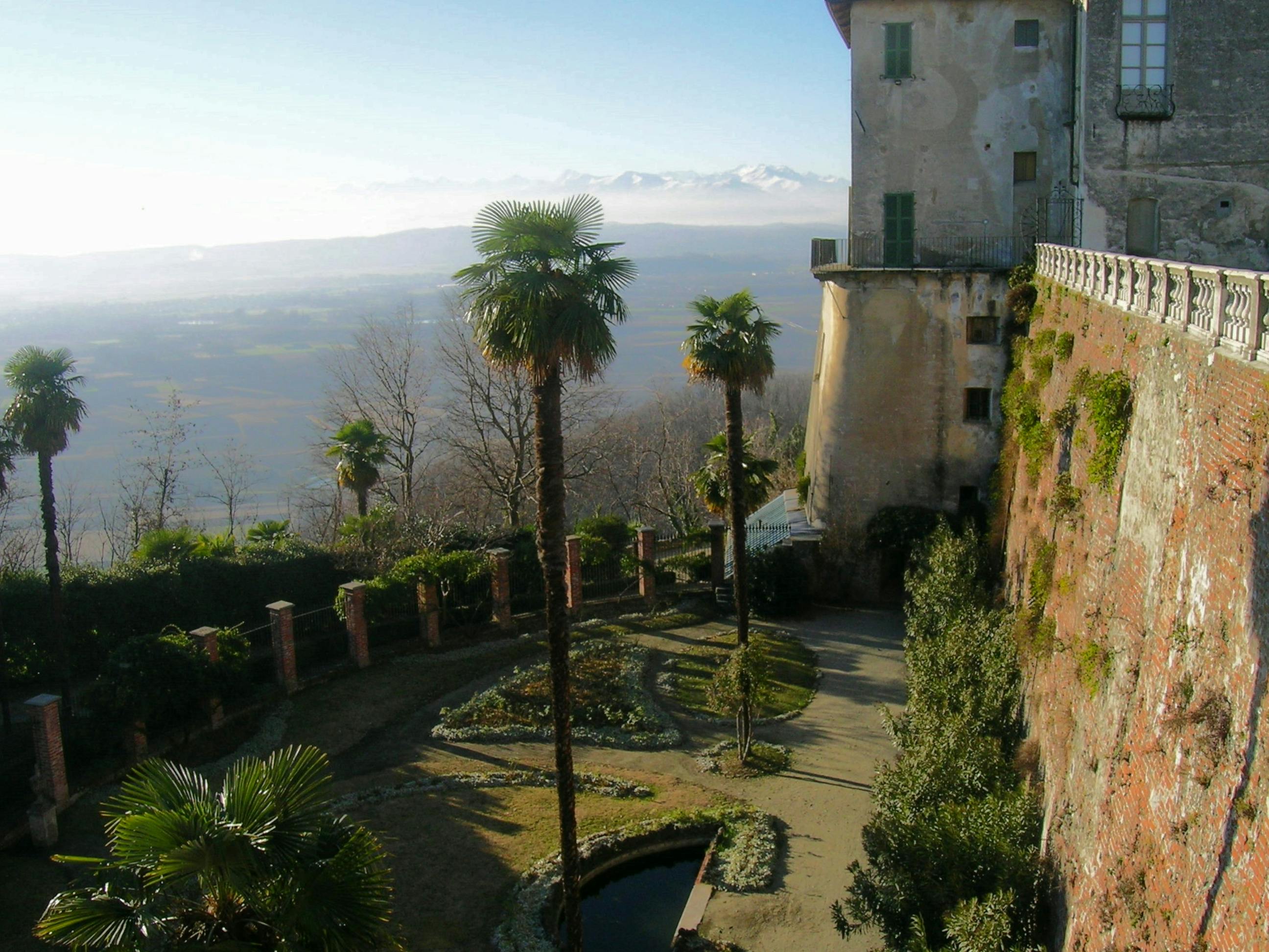 A garden with palm trees and a small pond beside an old stone building overlooking a vast landscape with distant mountains.