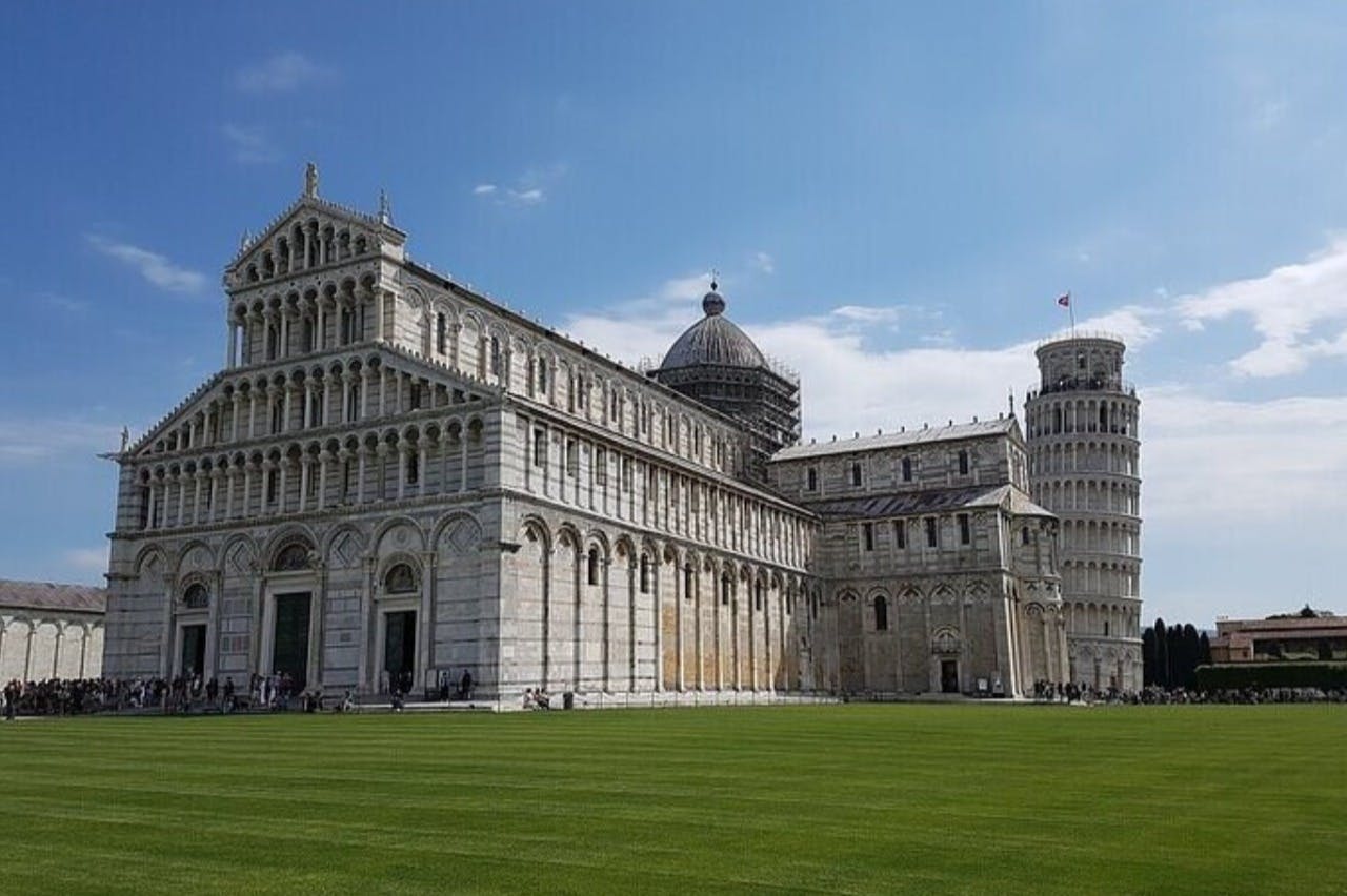 Ένας περίπλοκος καθεδρικός ναός στέκεται δίπλα σε ένα θολωτό κτίριο και στον Πύργο της Πίζας μια ηλιόλουστη μέρα, σε ένα καταπράσινο γκαζόν.