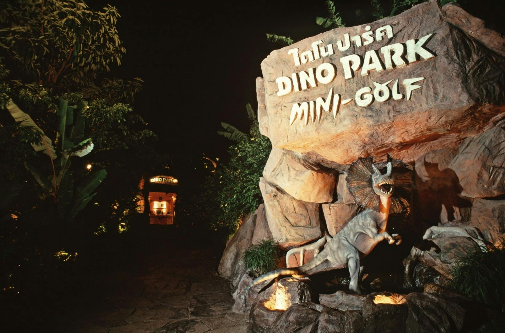 Wstęp do parku mini-golfa w nocy, z posągiem dinozaura i podświetlaną tablicą z napisem „Dino Park Mini-Golf”.