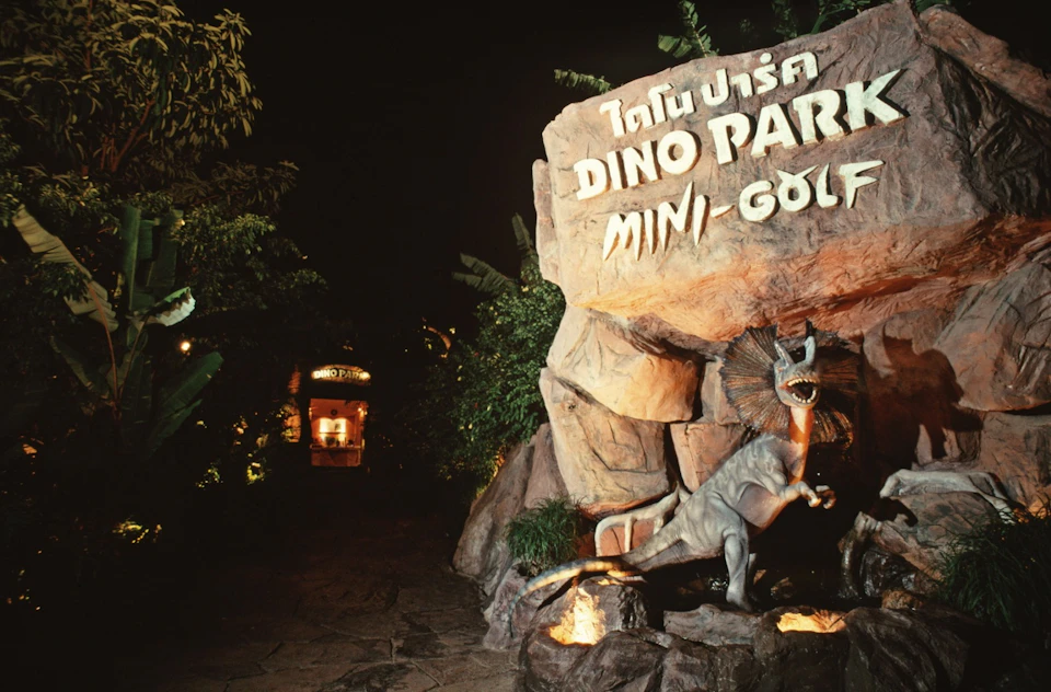 Entrance to Dino Park Mini-Golf at night, featuring a dinosaur statue, illuminated signs, and lush vegetation.