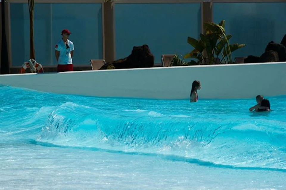 A person swims in a pool with waves, while another stands nearby in a lifeguard uniform. Tropical plants are visible in the background.