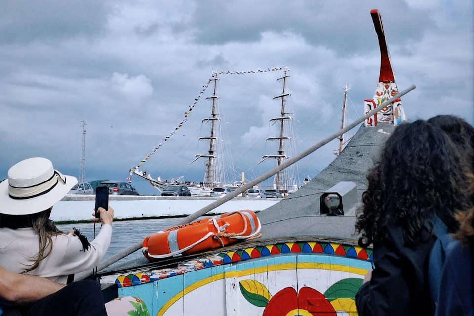 A person photographs a docked tall ship with decorative flags. The foreground has a colorful boat with a lifebuoy on its side.