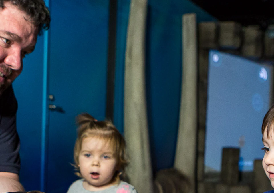 A man shows a turtle to three children in a room with a blue wall and wood-like pillars in the background.