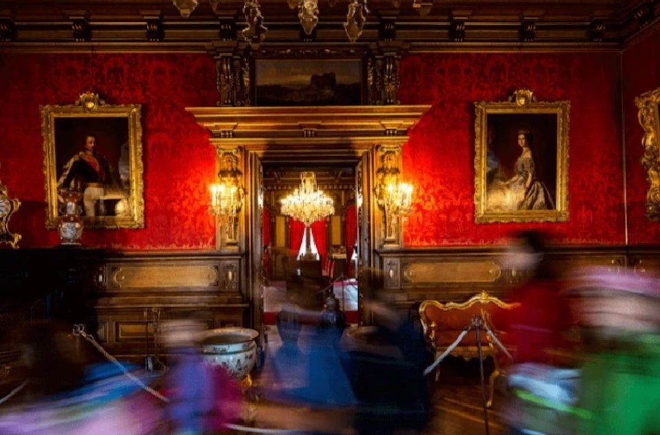 Lavish room with red walls, ornate decorations, chandeliers, and portraits. Blurred figures in the foreground suggest movement.
