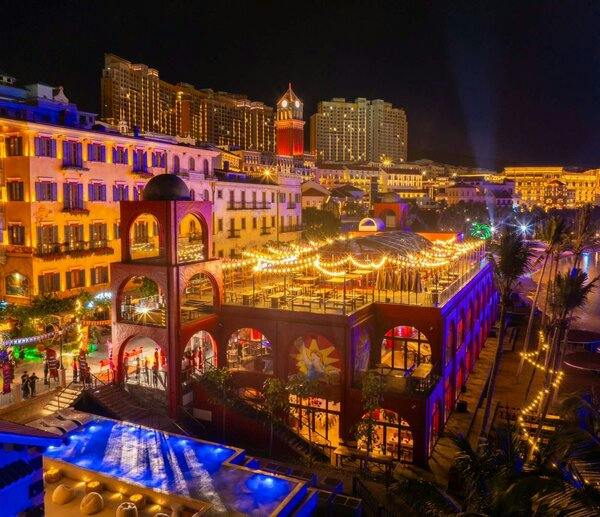 A lively nighttime cityscape with illuminated buildings, a vibrant plaza, and a pool in the foreground.