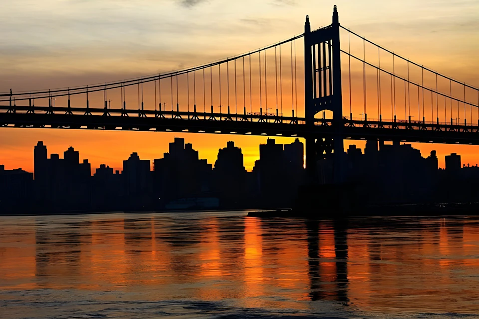 A suspension bridge silhouetted against a vibrant orange and yellow sunset with a city skyline and water below.