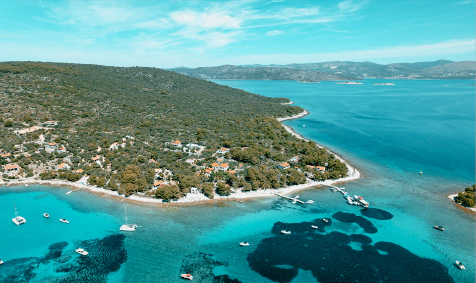Aerial view of a wooded coastal area with scattered houses, surrounded by clear turquoise water, boats, and distant mountains.