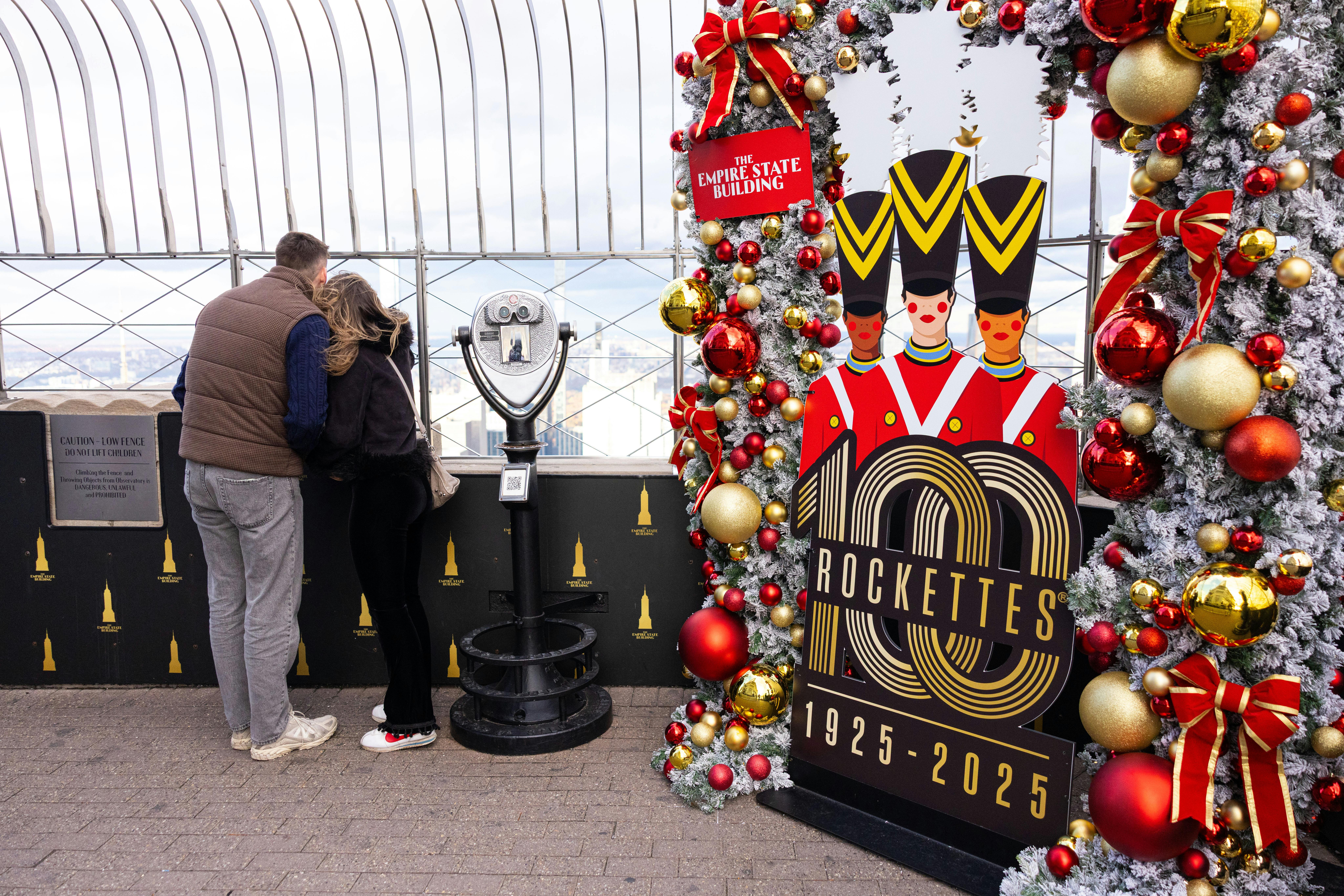 To personer står i nærheden af en møntbetjent seer på Empire State Buildings observationsdæk, dekoreret med elementer med ferie-tema, herunder et "Rockettes 100 Years "-skilt og festlige ornamenter.