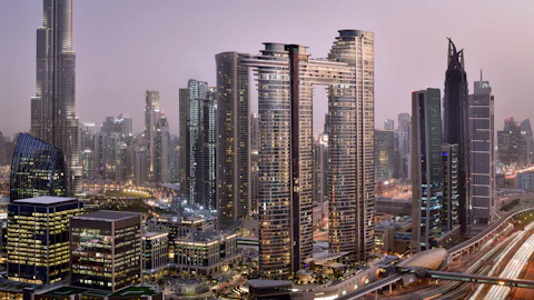 Panoramic view of a city skyline at dusk with modern skyscrapers, illuminated office buildings, and busy roads.