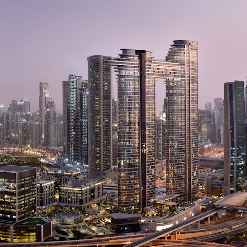 Panoramic view of a city skyline at dusk with modern skyscrapers, illuminated office buildings, and busy roads.