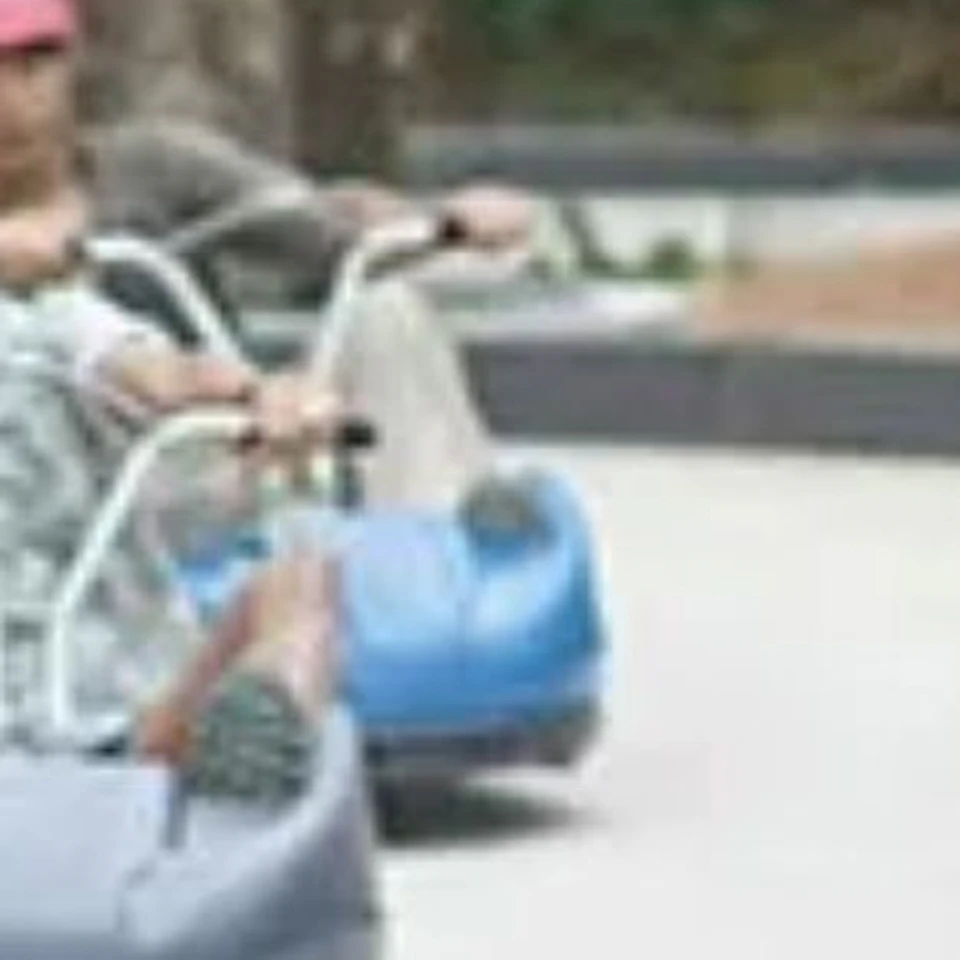People riding down a paved track on small, colorful luge carts with helmets in a lush, forested outdoor setting.