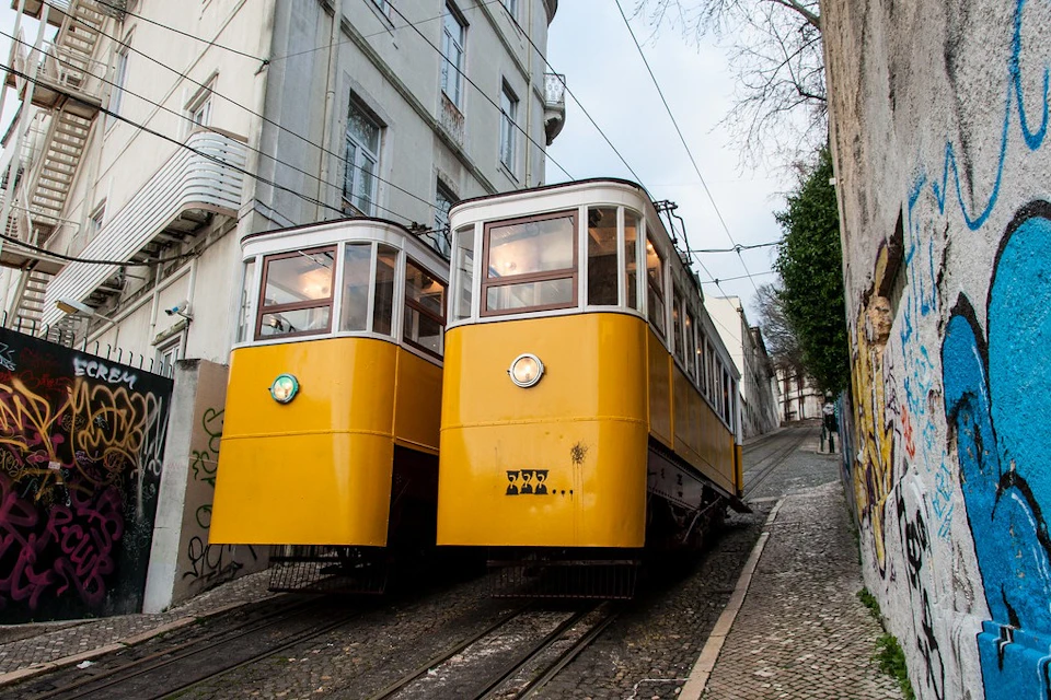 Two yellow trams on parallel tracks move up a steep, cobblestone street between buildings with graffiti on the walls.