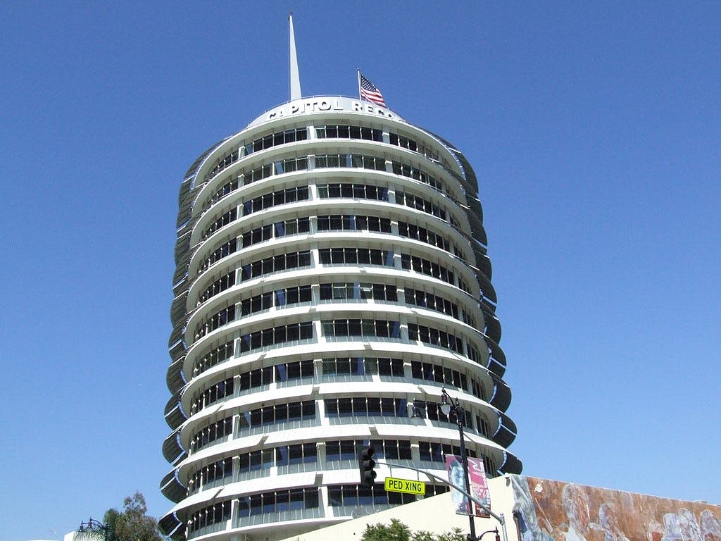 Un grand bâtiment cylindrique avec plusieurs balcons ronds, portant l'inscription "CAPITOL RECORDS", un drapeau américain au sommet et un ciel bleu.