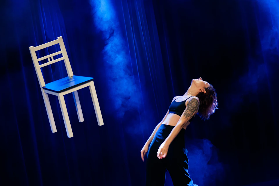 A person in a black outfit leans back dramatically under blue stage lighting, with a white chair suspended nearby.