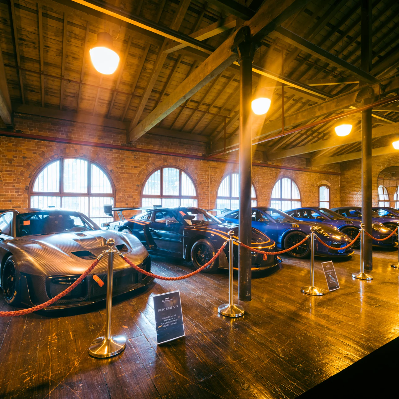 A row of luxury cars displayed in a warmly lit brick-walled showroom with wooden beams and roped-off sections.