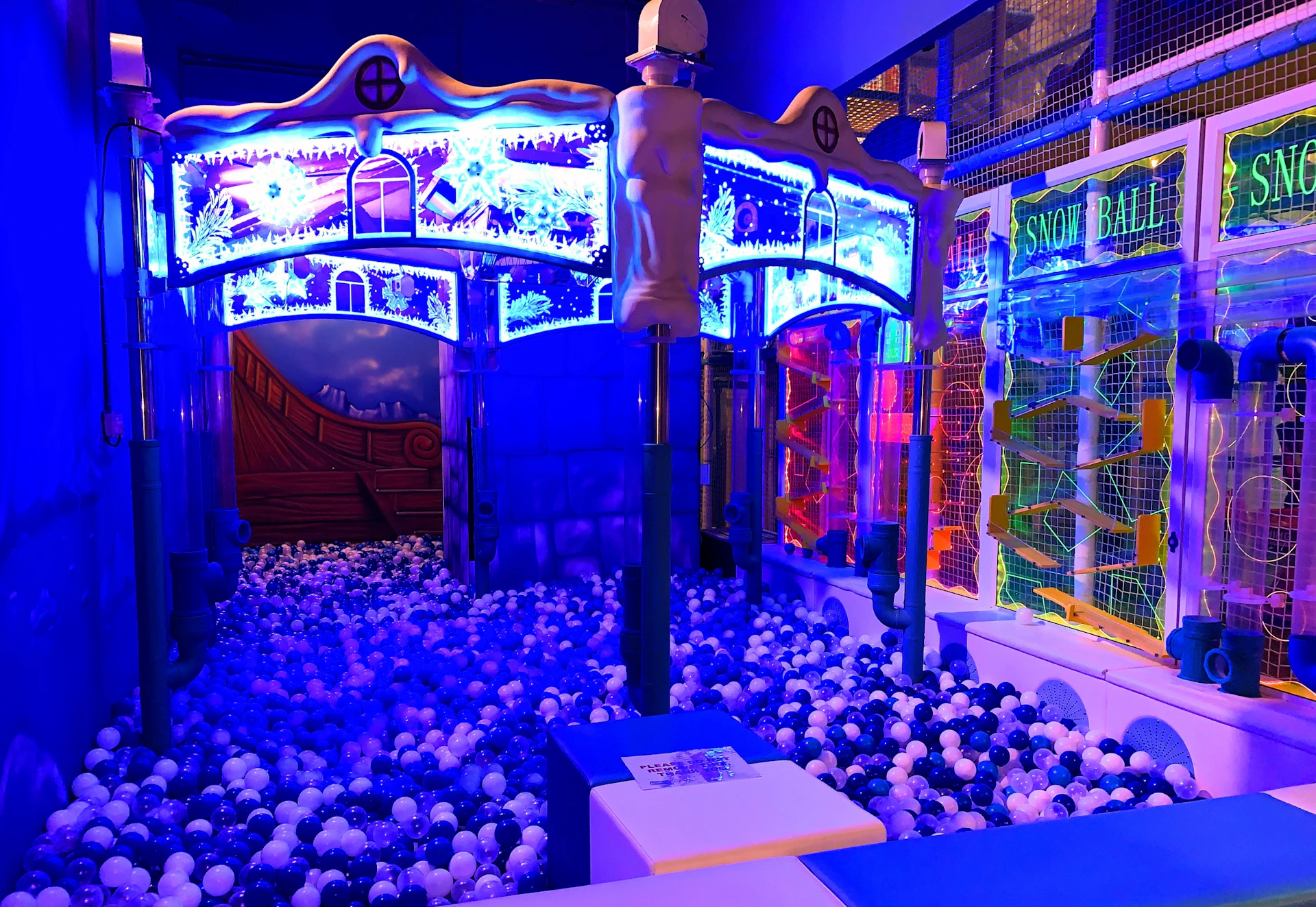 A colorful indoor playground with a ball pit, illuminated with blue and purple lights and themed decorations.