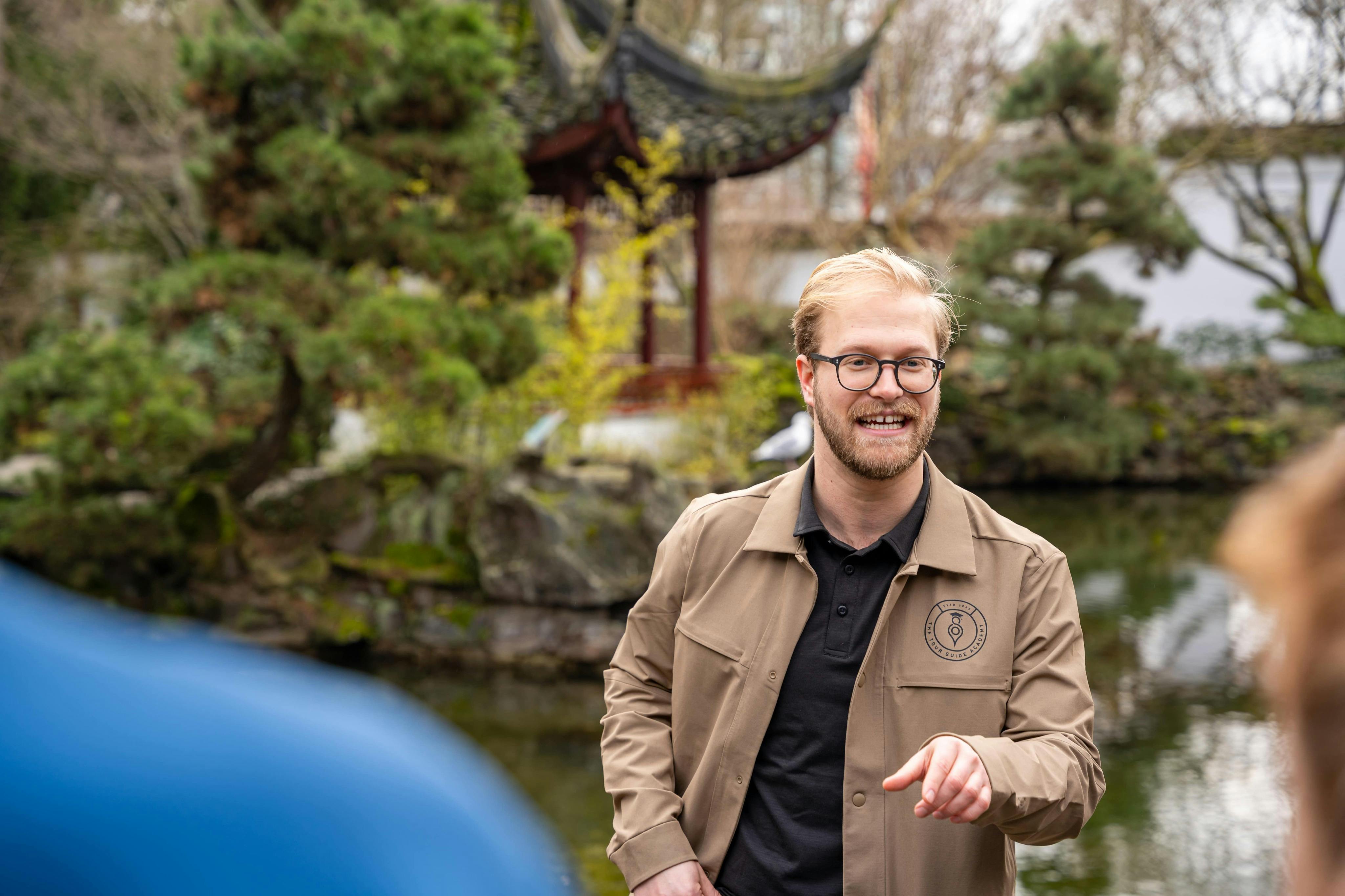 Muž v opáleném saku a brýlích stojící v zahradě s rybníkem a tradičním pavilonem v pozadí.