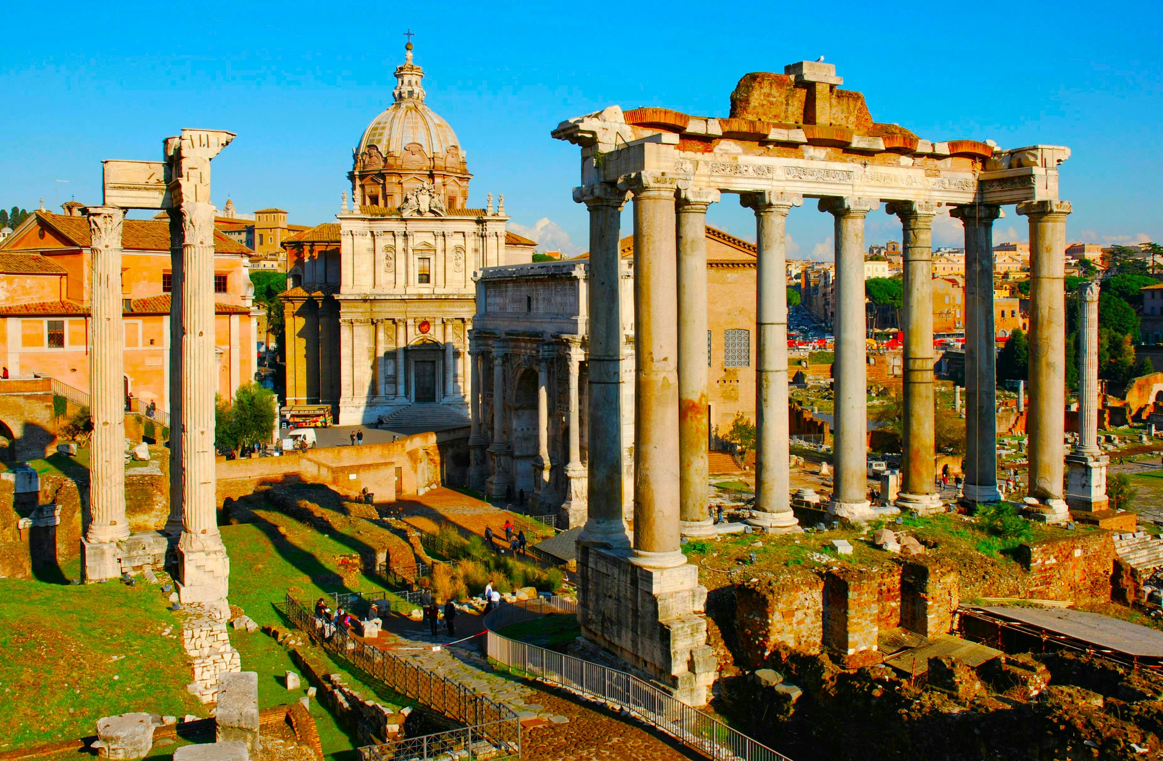 Ruins of ancient Roman columns and structures under a blue sky, with a dome and surrounding buildings in the background.
