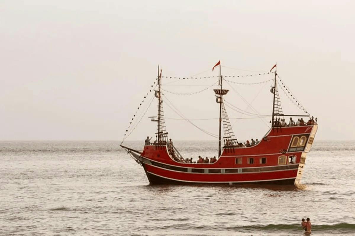 A red tourist ship with decorative lights and flags sails on calm ocean waters under a cloudy sky.