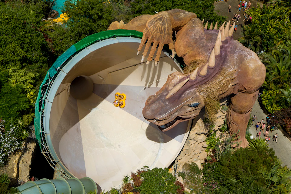 Two people riding a yellow raft slide inside a large funnel with a dragon sculpture wrapping around it. Trees surround the area.