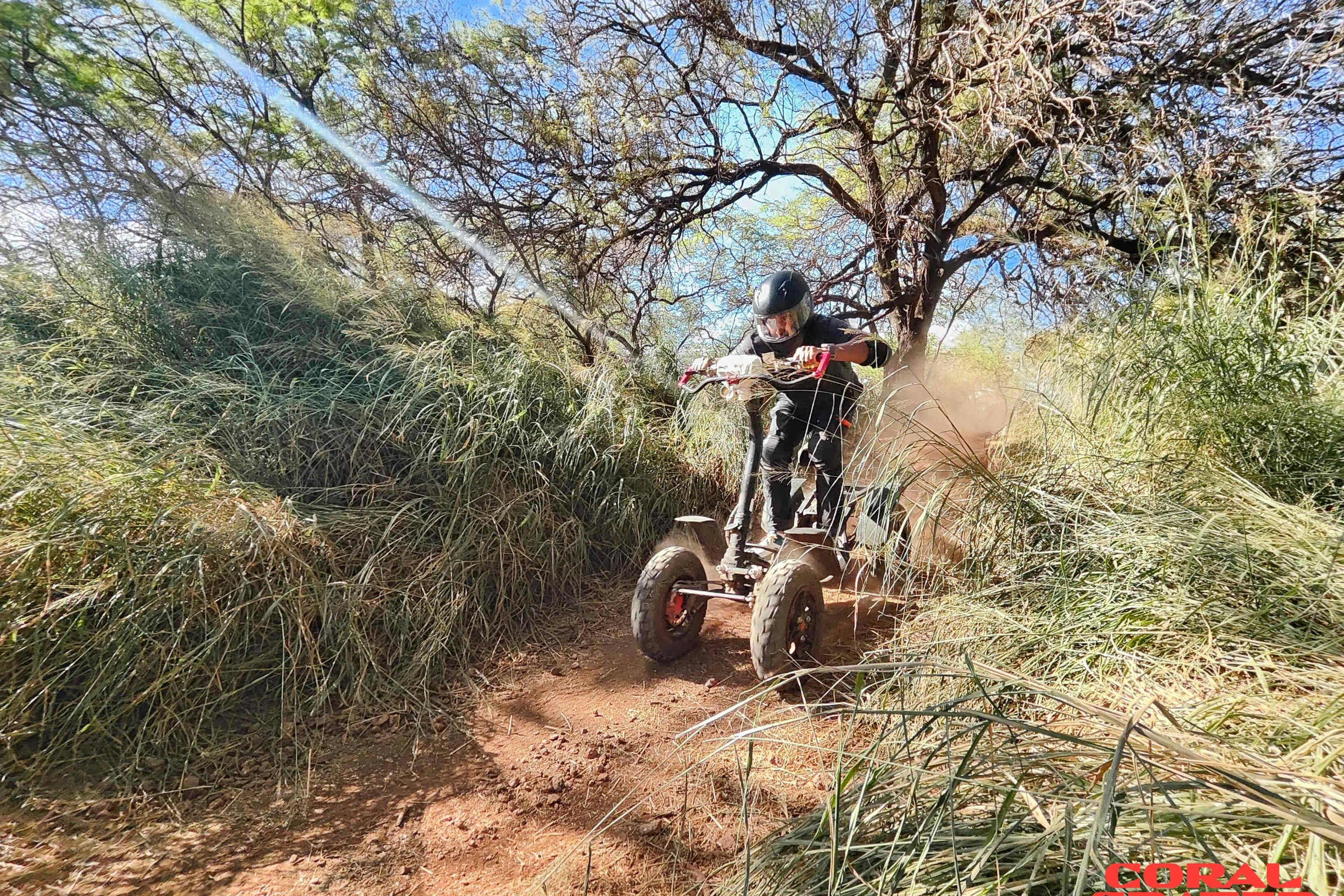 Osoba jedoucí v terénním vozidle po špinavé cestě hustou trávou a stromy, která nakopává prach. Jasný, slunečný den.