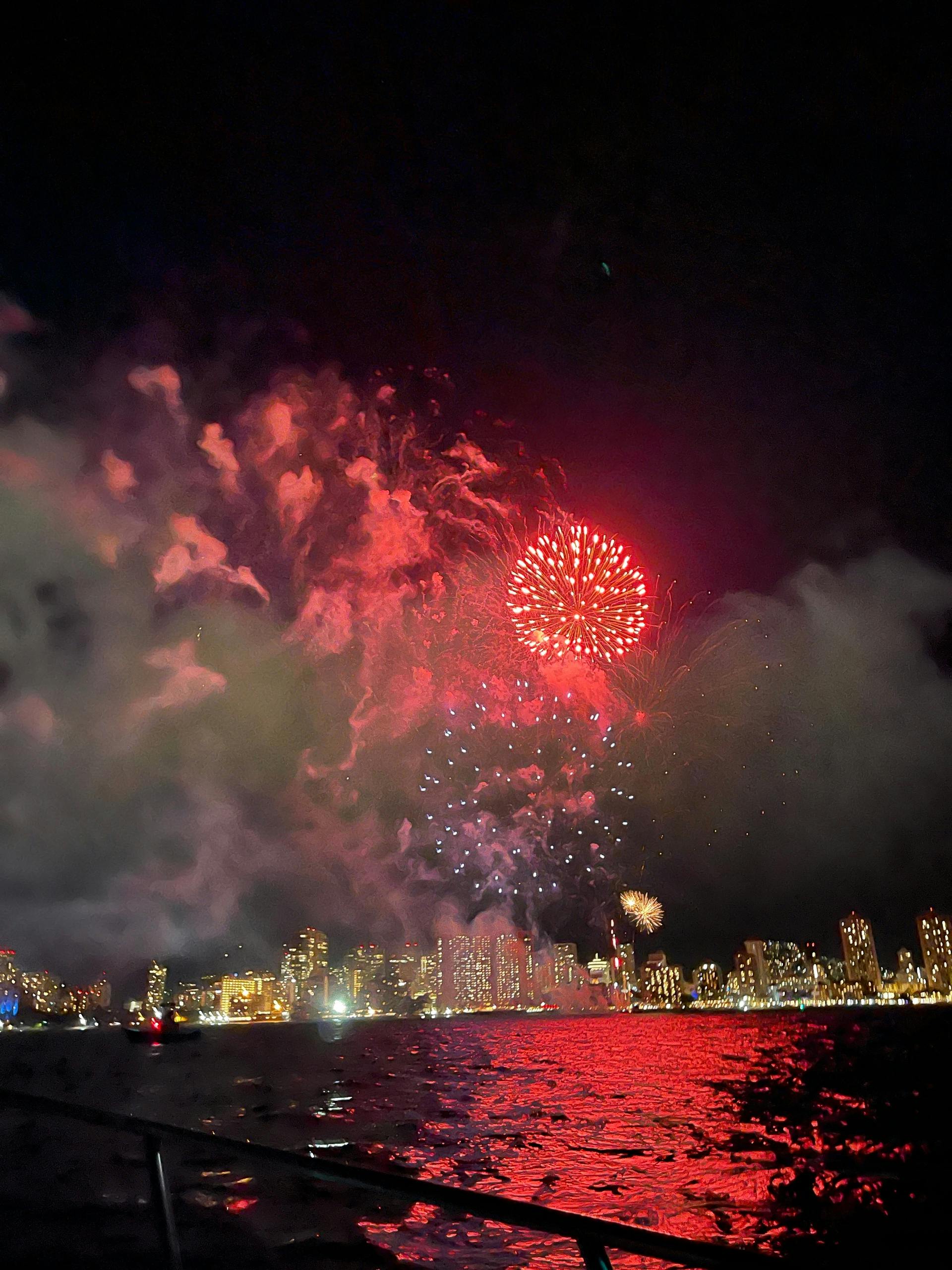 Ein helles Feuerwerk, das den Nachthimmel über einer beleuchteten Stadtsilhouette erhellt, mit Reflexionen auf dem Wasser darunter.