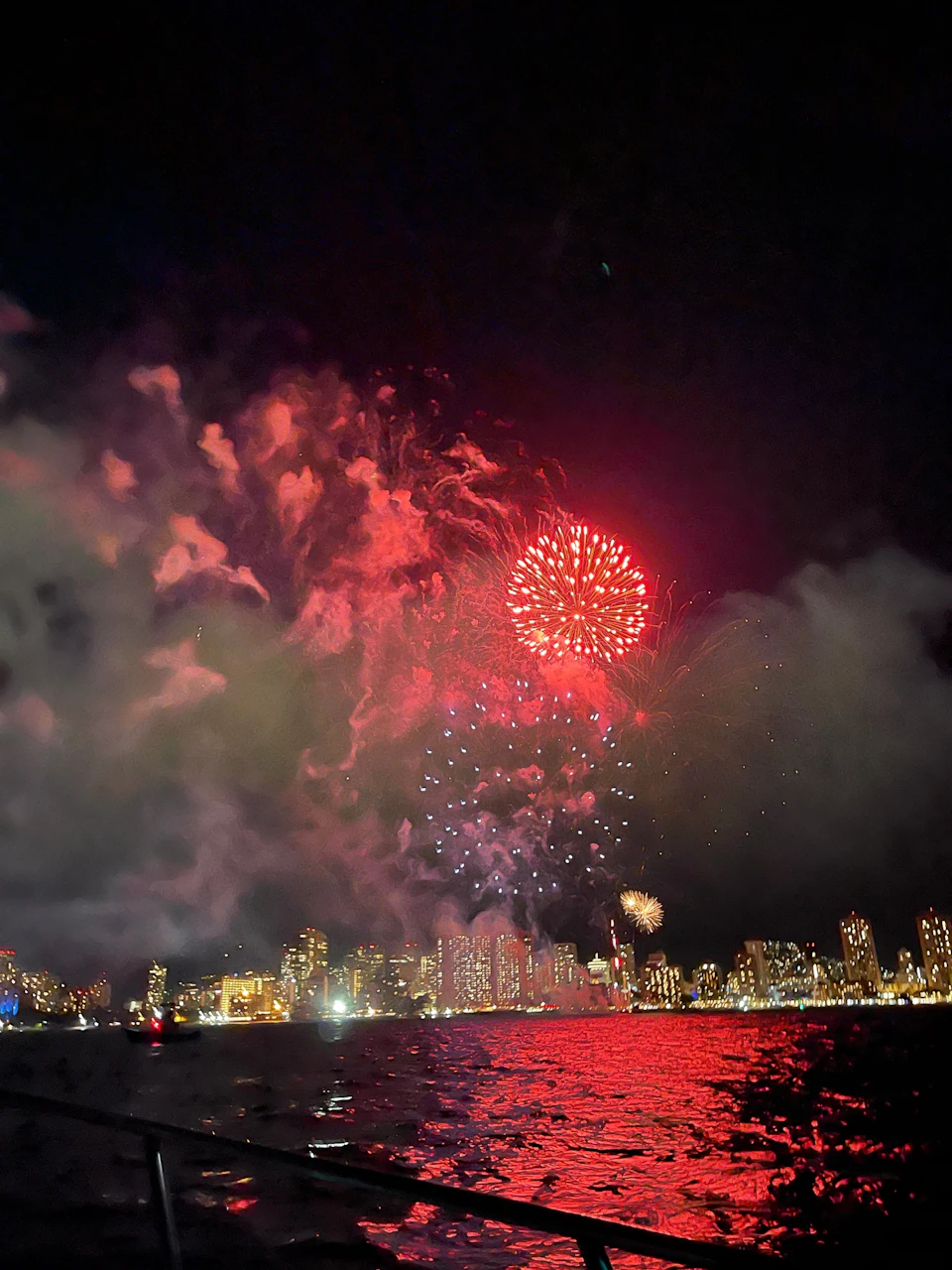 Bright fireworks light up the night sky above a cityscape, with reflections visible on the water below.