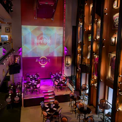En Hard Rock Café-interiör med bord, scen, memorabilia på väggar och en stor "Hard Rock Café" -logotypprojektion.