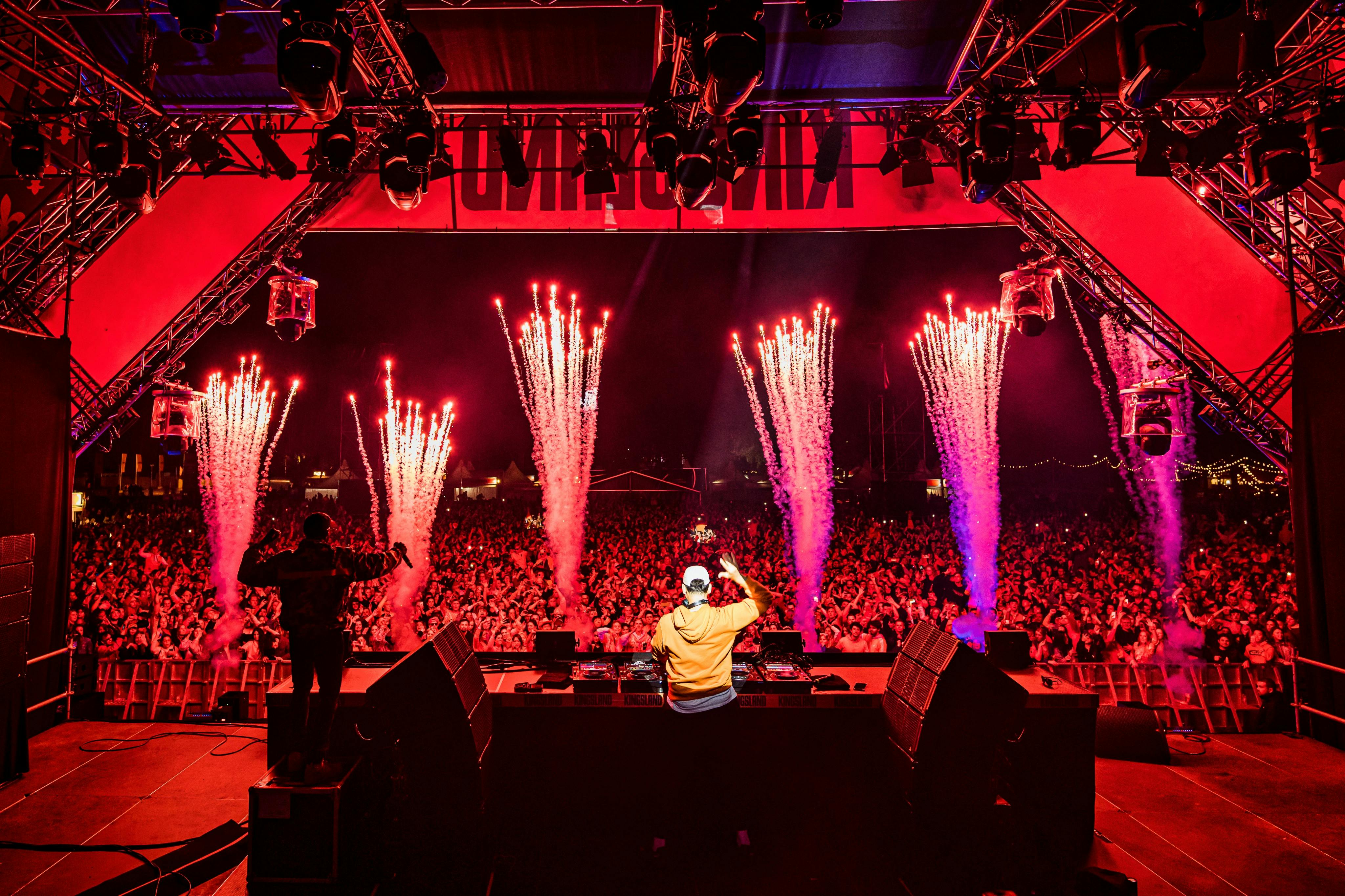 En DJ uppträder på scenen på en musikfestival, inför en stor, energisk publik. Fyrverkerier brast i bakgrunden.