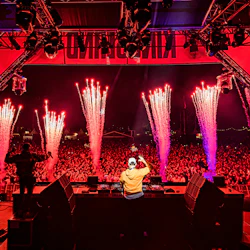 En DJ uppträder på scenen på en musikfestival, inför en stor, energisk publik. Fyrverkerier brast i bakgrunden.